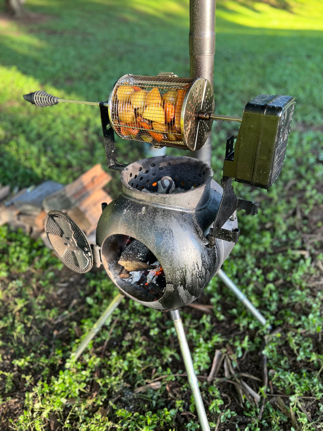 The Ozpig Rotisserie Basket Small from Ozpig is filled with sliced potatoes, roasting over a compact outdoor wood stove on a tripod, with sunlight and grass in the background—a must-have accessory for rotisserie cooking outdoors.