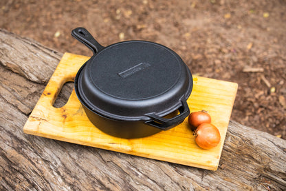 The Campfire Cast Iron Combo Cooker 3.2 Quart with a lid rests on a wooden board beside two brown onions, all set on a rustic outdoor wooden surface.