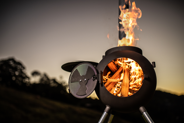 The Ozpig Series 2 Portable Wood Fire Stove by Ozpig is a round, black metal outdoor cooker on three legs, its open door revealing a bright fire—perfect for backyard entertaining against a dusk sky and grassy landscape.