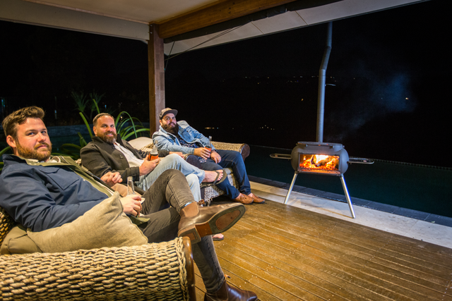 Three men relax at night on wicker furniture under a covered deck beside the Ozpig Big Pig by Ozpig, a wood-burning outdoor stove. Flames glow warmly, making it a cozy heating solution against the dark backdrop with distant lights.