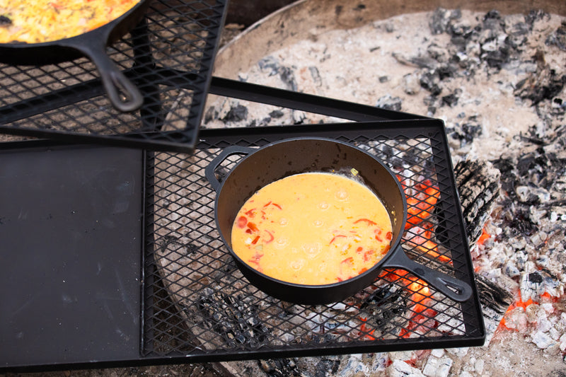 The Campfire Cast Iron Combo Cooker 3.2 Quart cooks a yellow and red egg mixture over an open campfire on a metal grate, with glowing embers and ashes visible beneath.