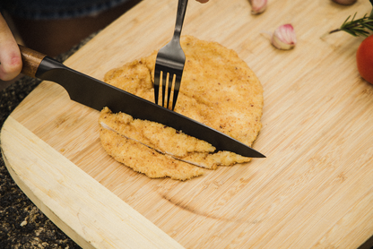 A hand slices a breaded chicken cutlet with a stainless steel knife and fork from the Campfire 3pc Premium Knife Set on a wooden cutting board, with garlic cloves, tomato, and rosemary in the background.