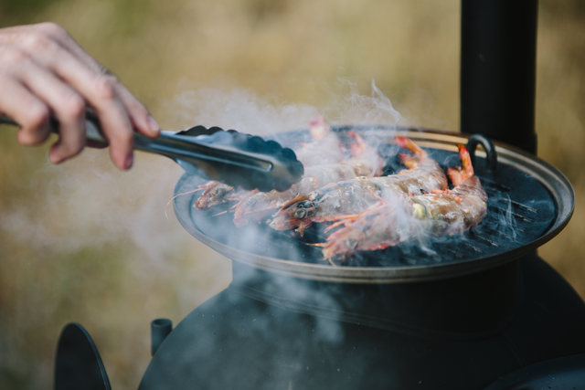 A hand uses tongs to grill prawns over an Ozpig Series 2 Portable Wood Fire Stove by Ozpig, with smoke rising from the seafood. The blurred background emphasizes the vibrant grilling scene—ideal for backyard entertaining.