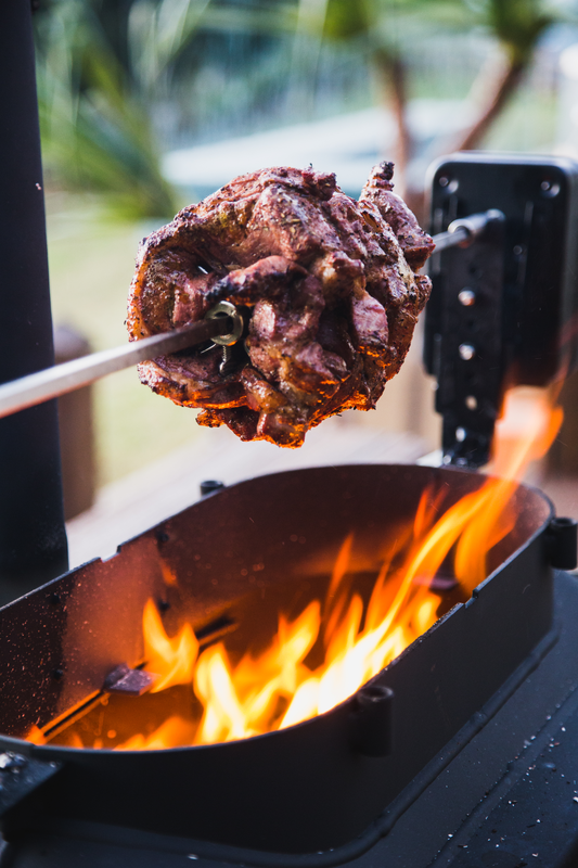 The Ozpig Big Pig Rotisserie Kit by Ozpig slowly roasts seasoned meat over an open wood fire, while blurred greenery appears in the background for an authentic outdoor cooking experience.
