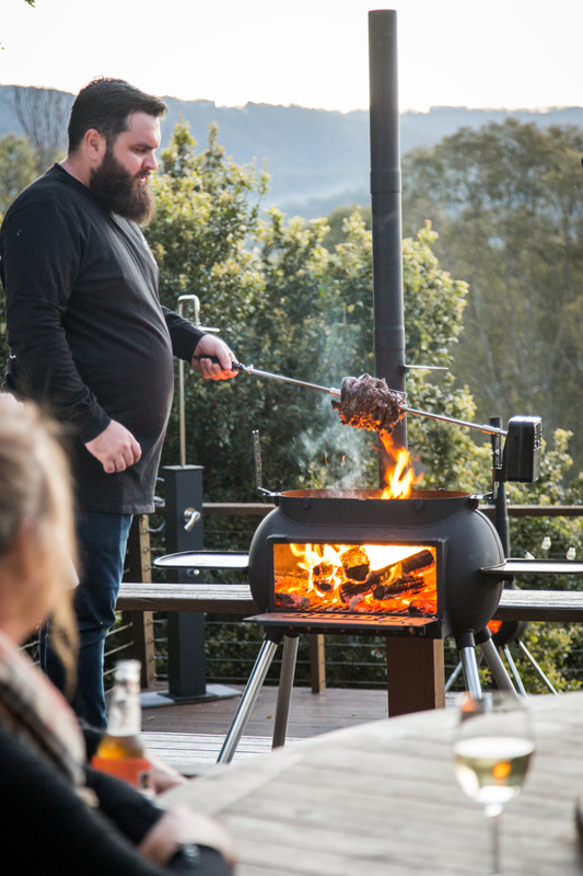 A man grills meat over an outdoor firepit using the Ozpig Big Pig Rotisserie Kit, while two people sit at a table with drinks, surrounded by trees and hills in the background.