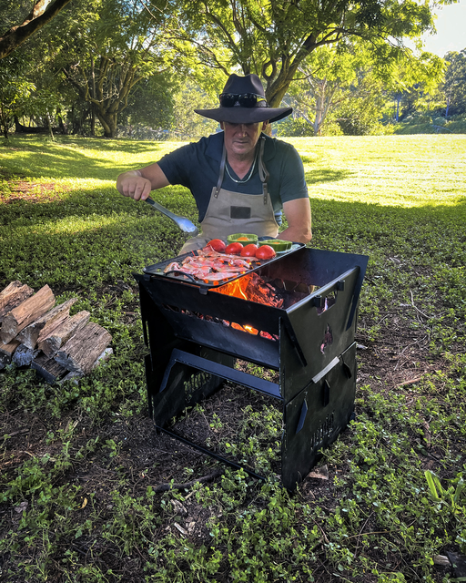 Grilling meat and veggies outdoors with the Ozpig 3-in-1 Flat Pack Fire Pit, a person in a wide-brim hat and apron cooks over an open flame, surrounded by grass and trees, with a stack of firewood nearby.