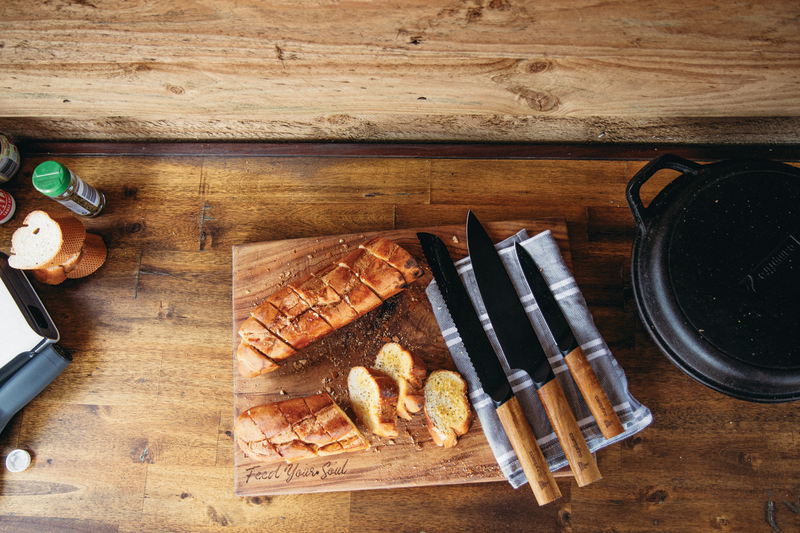 Sliced bread and two herb-topped slices sit on a wooden cutting board with a cast iron pot nearby, while the Campfire 3pc Premium Knife Set rests on a kitchen towel atop the wooden countertop.