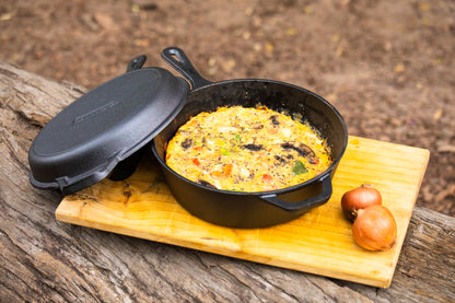 A Campfire Cast Iron Combo Cooker 3.2 Quart, pre-seasoned, holds a baked egg dish on a wooden cutting board outdoors. The cooker's lid is nearby, with two whole onions beside the skillet.