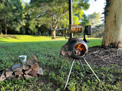 The Ozpig Rotisserie Basket Small by Ozpig holds a chicken above the flames of an outdoor stove, roasting on grass beside firewood, with trees and lush greenery in the background.