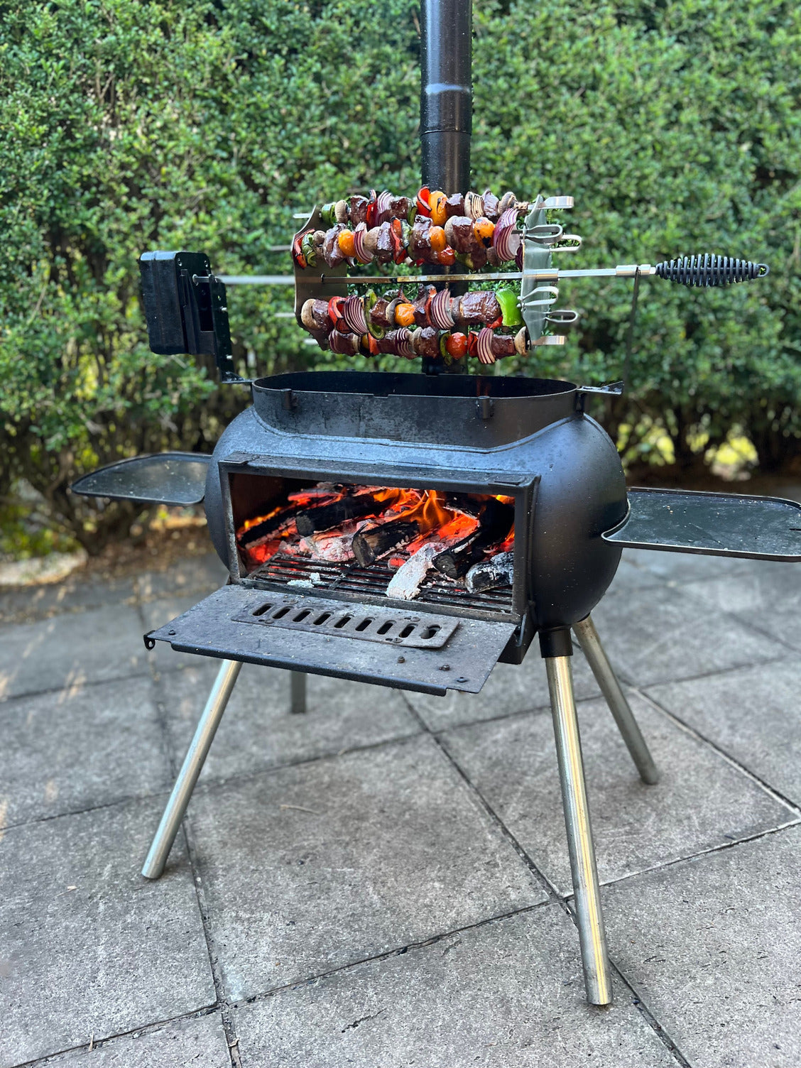 The Ozpig Kebab Rotisserie Set by Ozpig is shown in black, with stainless steel skewers of meat and veggies rotating over burning wood and charcoal on a tiled patio, green bushes in the background.