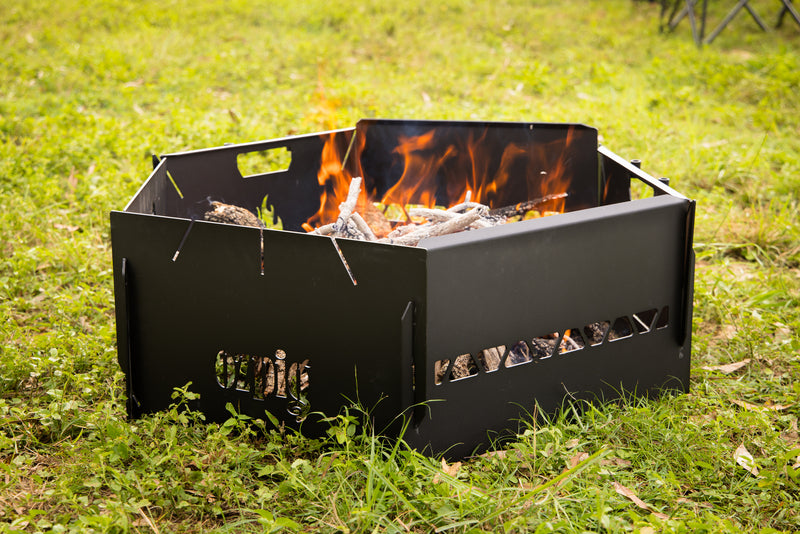 The Ozpig 3-in-1 Flat Pack Fire Pit, a black metal hexagonal fire pit with burning wood and flames, sits on green grass outdoors.