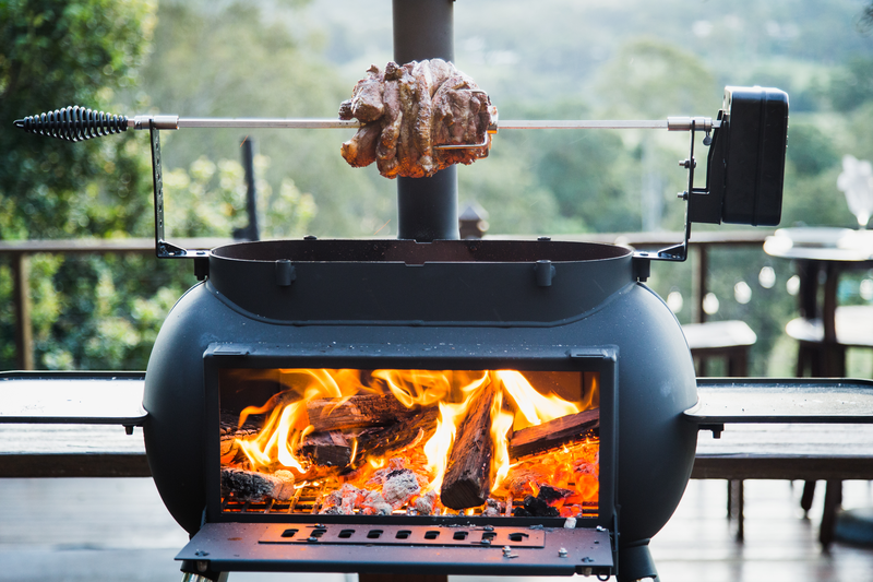 A large cut of meat roasts over an open fire with the Ozpig Big Pig Rotisserie Kit by Ozpig, set inside a black outdoor grill on a wooden deck surrounded by trees and greenery.