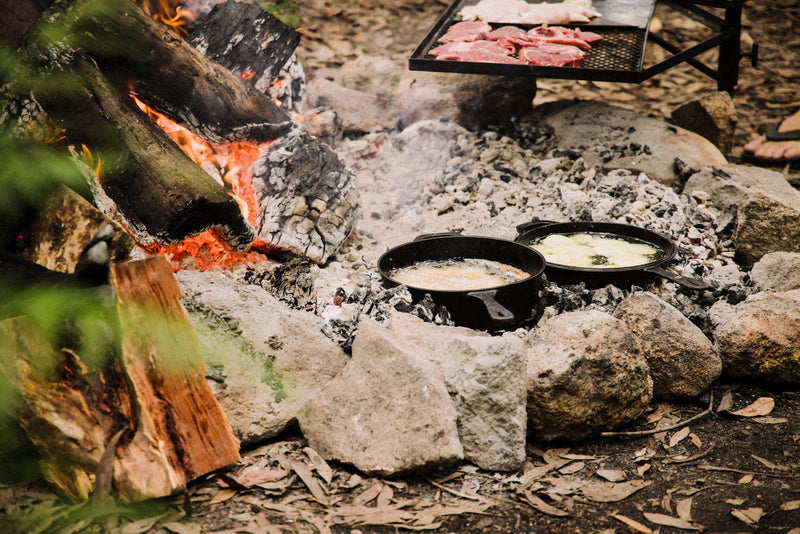 Two Campfire Cast Iron Combo Cooker 3.2 Quart pans cook over hot coals inside a stone fire ring, while a grill holds meat above a burning log fire—perfect for camp oven meals in the woods.