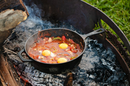 The Campfire Cast Iron Combo Cooker 3.2 Quart is shown cooking eggs, tomatoes, and vegetables over a campfire with charred wood, ash, and green grass in the background.