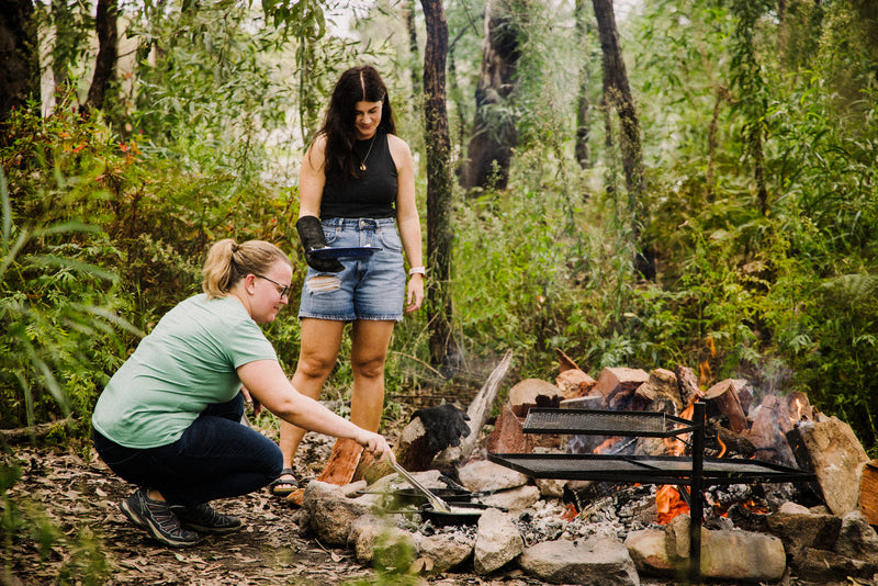Two people are cooking over an open campfire in a forest using the Campfire Cast Iron Combo Cooker 3.2 Quart. One person squats to handle the cooker above the flames while another watches with a plate, surrounded by rocks and logs beneath tall trees.