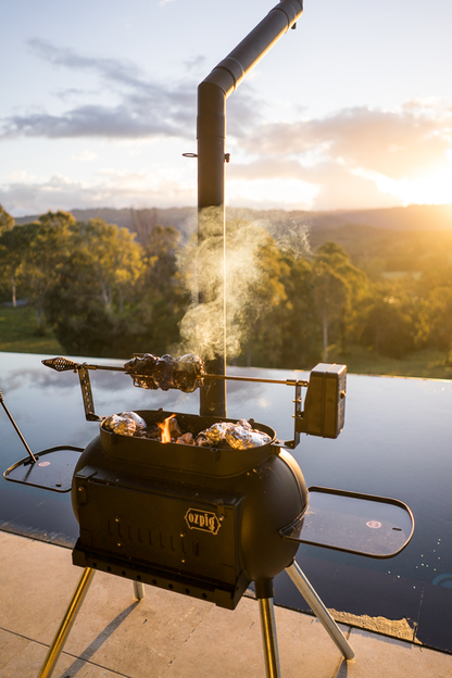 The Ozpig Big Pig Rotisserie Kit by Ozpig is grilling meat and foil-wrapped food beside a pool at sunset, with smoke rising against a scenic backdrop of trees and hills.