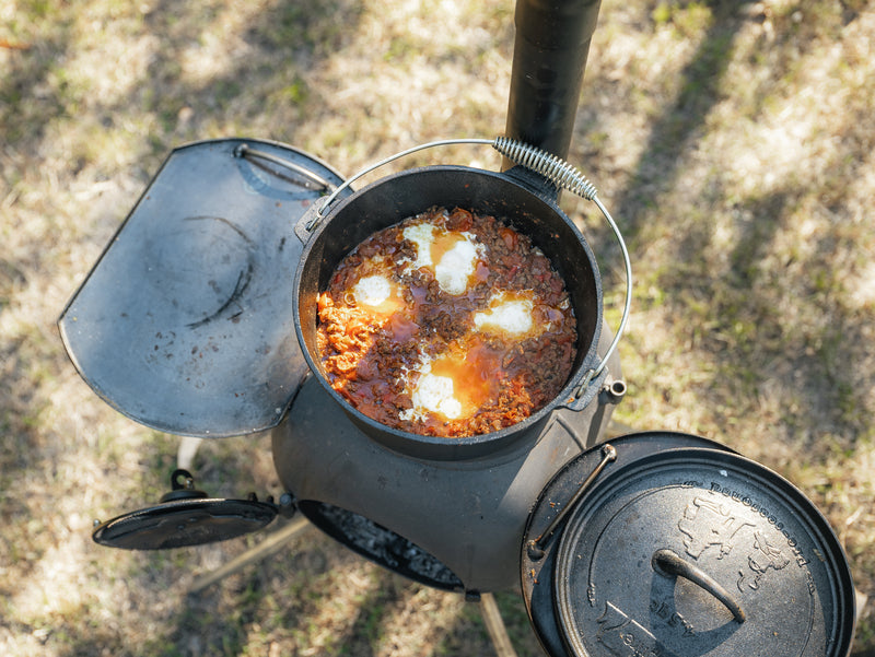The Campfire 4.5 Quart Camp Oven, filled with tomato stew and poached eggs, sits on an outdoor wood-burning stove as sunlight casts shadows on the grassy ground, capturing the spirit of outdoor cooking.