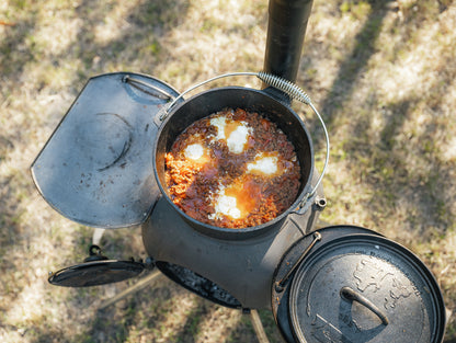 The Campfire 4.5 Quart Camp Oven, filled with tomato stew and poached eggs, sits on an outdoor wood-burning stove as sunlight casts shadows on the grassy ground, capturing the spirit of outdoor cooking.