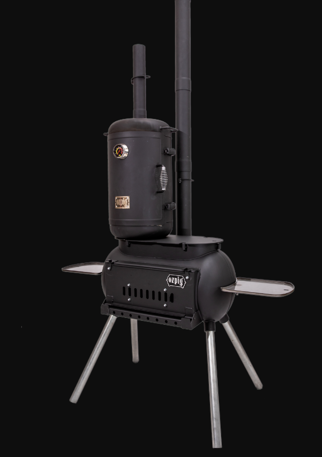 An Ozpig Big Pig Oven Smoker Adapter is paired with a black metal wood-burning stove featuring a vertical water heater attachment, two side shelves, and four angled legs, all set against a plain black background. The versatile Ozpig Big Pig model can be easily customized with the Oven Smoker Adapter for an enhanced outdoor cooking experience.