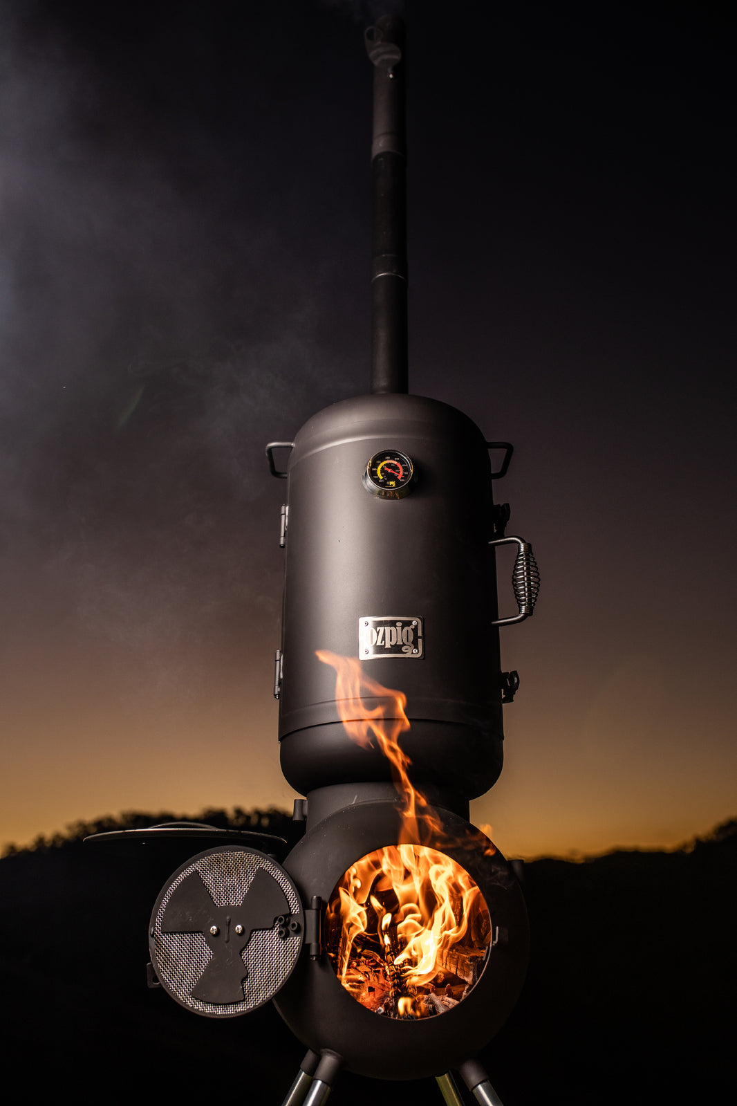 The Ozpig Oven Smoker by Ozpig is a black outdoor wood stove with a visible fire glowing inside, set against the backdrop of a dark sunset sky. Designed for outdoor cooking, it features a tall chimney and a front-mounted thermometer, making it perfect for achieving that authentic woodfired flavour.