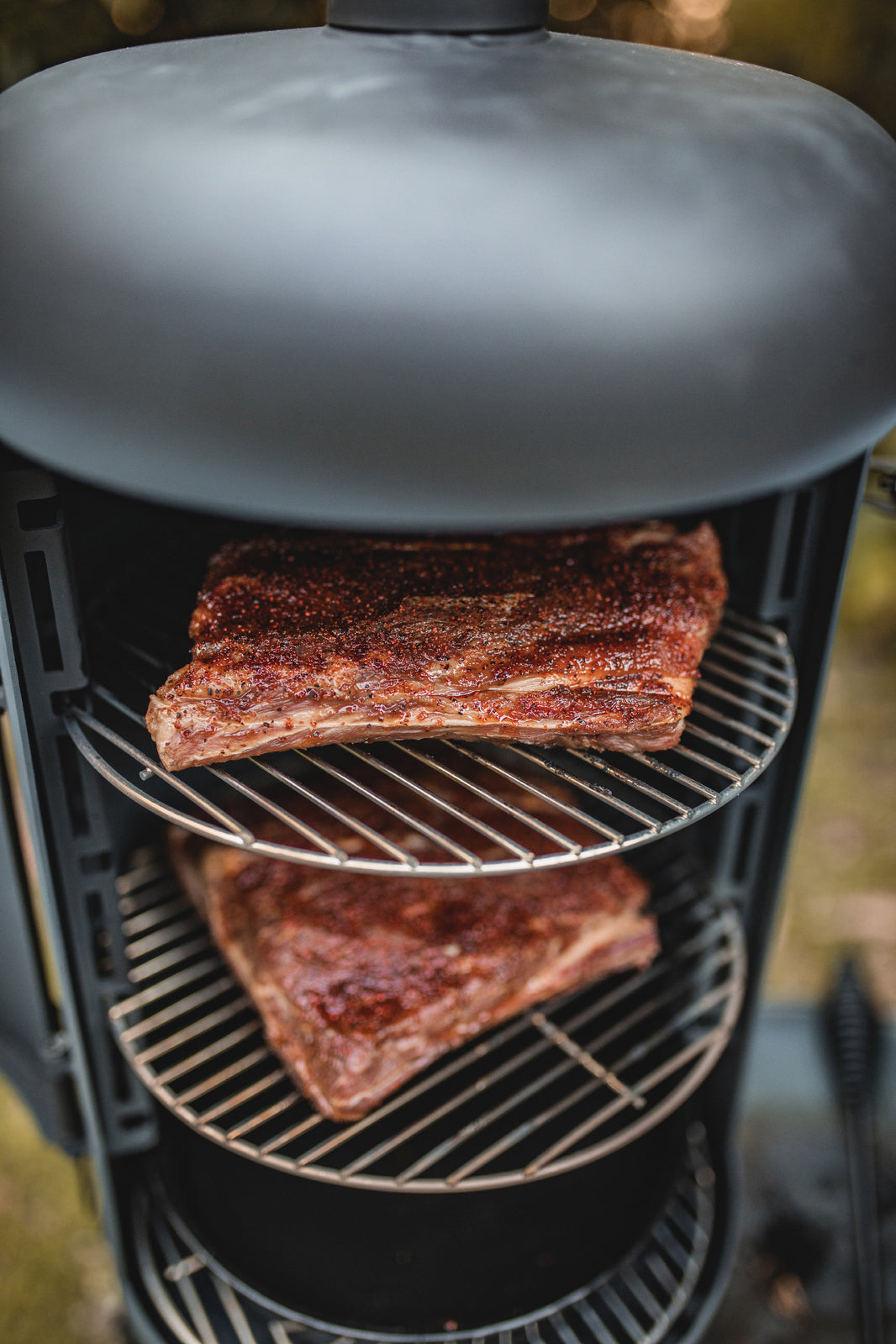 Two racks of seasoned meat smoke on separate shelves inside the Ozpig Oven Smoker by Ozpig, with the partially visible lid at the top and a blurred outdoor background highlighting the woodfired flavour of authentic outdoor cooking.