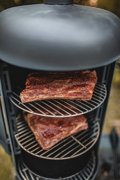 Two racks of seasoned meat smoke on separate shelves inside the Ozpig Oven Smoker by Ozpig, with the partially visible lid at the top and a blurred outdoor background highlighting the woodfired flavour of authentic outdoor cooking.