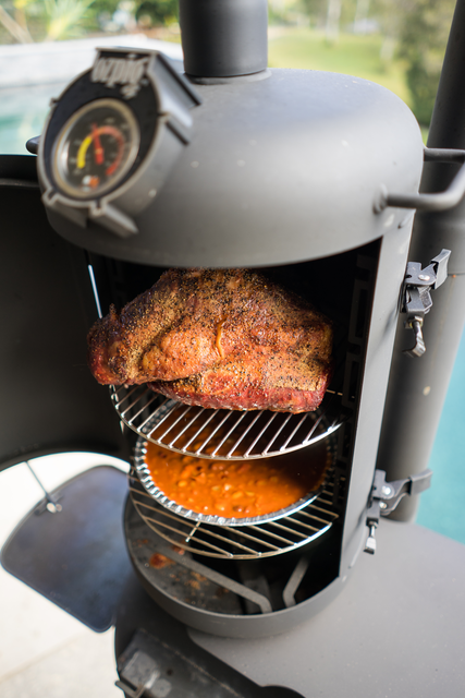 A seasoned brisket cooks on the top rack of the Ozpig Oven Smoker by Ozpig, absorbing rich woodfired flavour as orange-red drippings collect below, all set outdoors for the ultimate outdoor cooking experience.