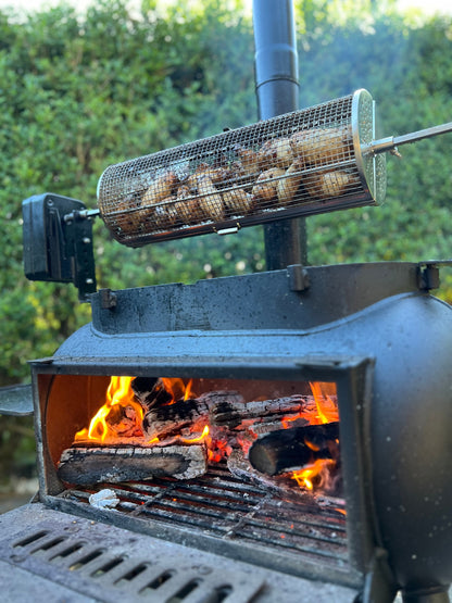 The Ozpig Rotisserie Basket Large by Ozpig is filled with mushrooms and cooks above an outdoor wood fire grill, with visible flames and burning logs below. Set against a green hedge background, this essential rotisserie accessory is perfect for elevating your wood-fired cooking experience.