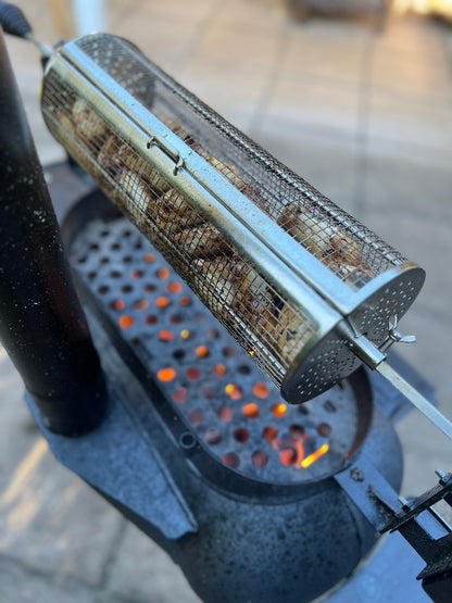 A metal mesh basket, like the Ozpig Rotisserie Basket Large by Ozpig, filled with peanuts is being roasted over a charcoal grill. Glowing coals are visible beneath, set against an outdoor tiled surface in the background—perfect for adding a touch of wood-fired cooking to your snack preparation.