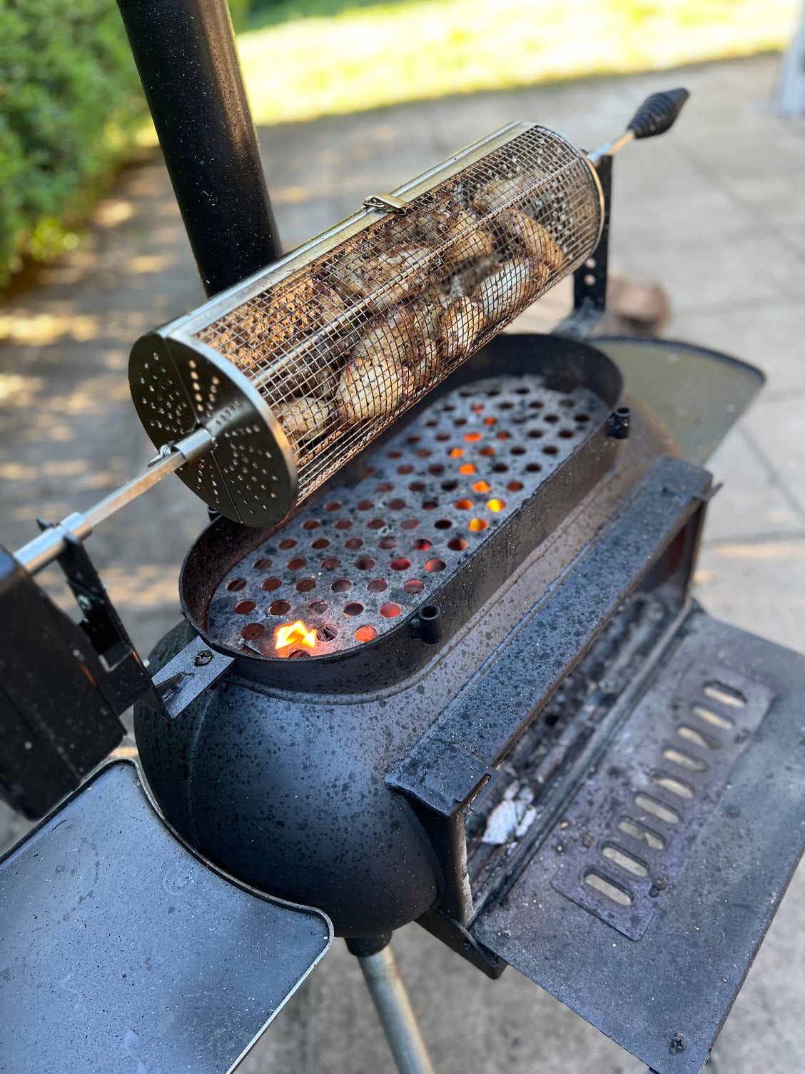 The Ozpig Rotisserie Basket Large by Ozpig, loaded with chicken, cooks over an open flame on a black outdoor grill, set on a patio surrounded by greenery—a perfect arrangement for authentic wood fired cooking.