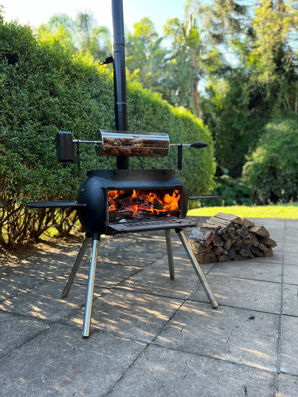 A black outdoor grill sits on a patio beside a stack of firewood, lush greenery in the background, with meat roasting over an open flame inside the Ozpig Rotisserie Basket Large by Ozpig—an ideal rotisserie accessory for wood fired cooking.