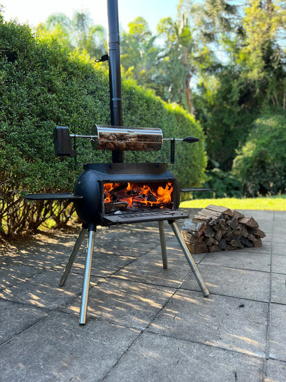A black outdoor grill sits on a patio beside a stack of firewood, lush greenery in the background, with meat roasting over an open flame inside the Ozpig Rotisserie Basket Large by Ozpig—an ideal rotisserie accessory for wood fired cooking.