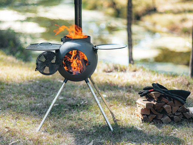 An Ozpig Series 2 Portable Wood Fire Stove by Ozpig, in black, stands on the grass with a fire glowing inside. Nearby, a stack of firewood and a pair of black gloves are placed—ideal for backyard entertaining. In the background, trees and water appear softly blurred.