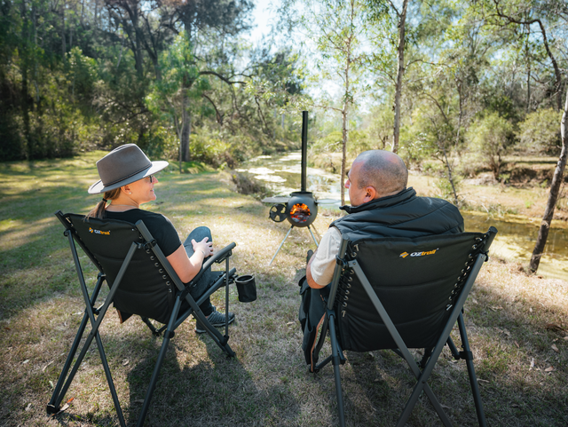 Two people sit in camping chairs by a creek in a forested area, talking and relaxing near an Ozpig Series 2 Portable Wood Fire Stove by Ozpig, with a fire burning, surrounded by trees and sunlight.