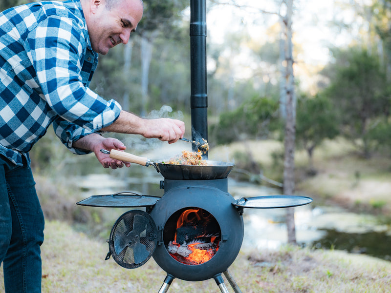 Wearing a plaid shirt, a man cooks food in a pan on the Ozpig Series 2 Portable Wood Fire Stove by Ozpig, with flames visible inside. Ideal for backyard entertaining, the scene is set against a backdrop of trees and a pond.