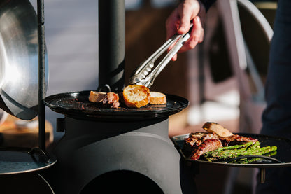 A person uses tongs to grill slices of bread on a black Ozpig Series 2 Portable Wood Fire Stove by Ozpig. Pieces of meat and asparagus rest on a tray beside the stove—perfect for backyard entertaining—as sunlight shines on the food and the cook’s hand.