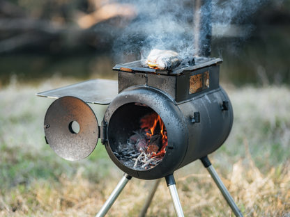 The Ozpig Traveller by Ozpig is a compact, black metal outdoor stove with its door open to reveal a lively fire inside. Fish sizzles on top as smoke drifts upward—an ideal companion for travel cooking and adventure—set outdoors on grass with a softly blurred background.