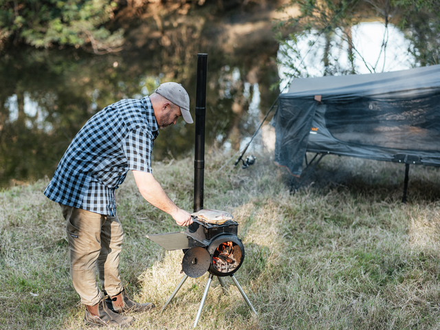 Wearing a plaid shirt and cap, a person cooks food outdoors on the Ozpig Traveller by Ozpig, near a tent and a creek amid trees and grass.