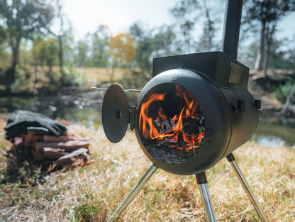 An Ozpig Traveller outdoor stove by Ozpig, crafted from black metal, stands on the grass near neatly stacked firewood and a river, surrounded by trees in daylight—a perfect adventure companion for travel cooking in the heart of nature.