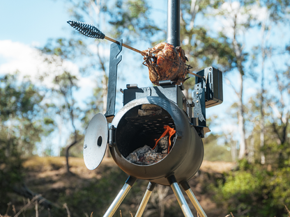 A whole chicken turns slowly over the fire inside a black Ozpig Traveller Rotisserie Kit—the ultimate portable charcoal rotisserie from Ozpig—set in an outdoor barrel smoker with a chimney, framed by trees and a clear blue sky.