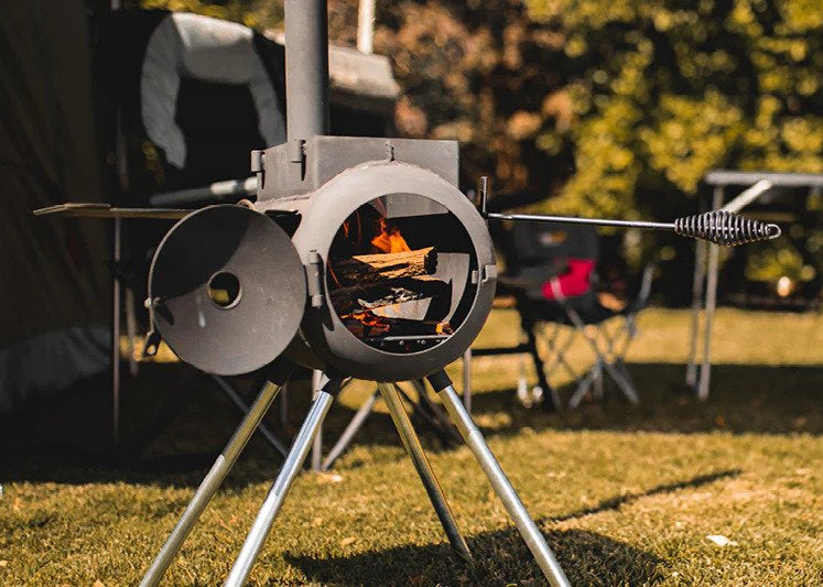 A portable metal wood stove with a round door open, showing burning logs inside, stands on grass with tents and camping chairs in the background on a sunny day.